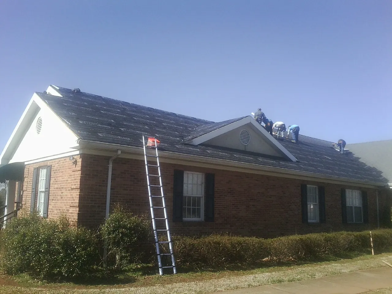 Crew tearing shingles off a brick ranch roof with a ladder set — Metal Roof Repair work by our crew in Olga