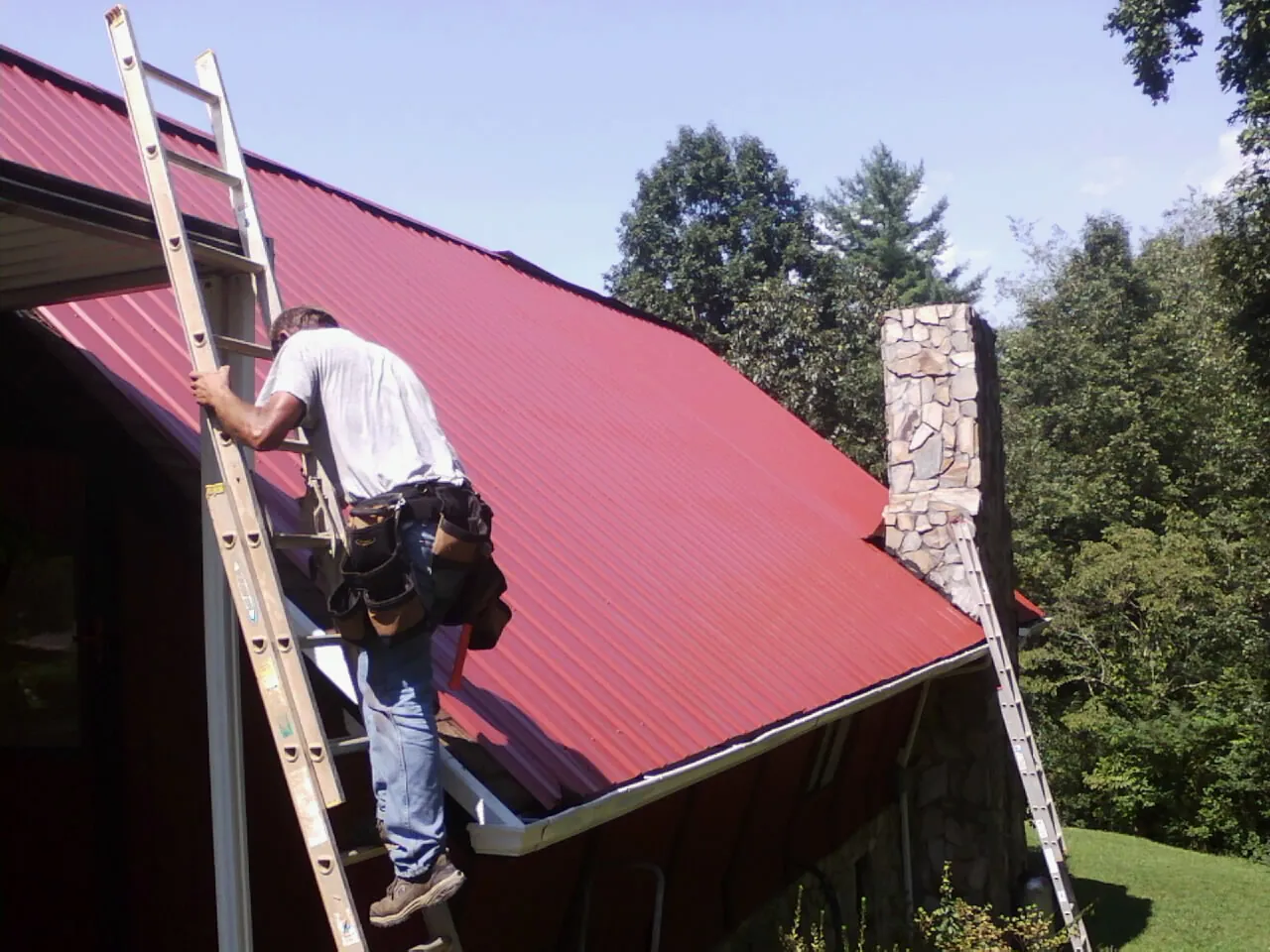 Roofer on a ladder beside a red metal roof and stone chimney — Soffit & Fascia Repair by Roofing Brothers in Olga